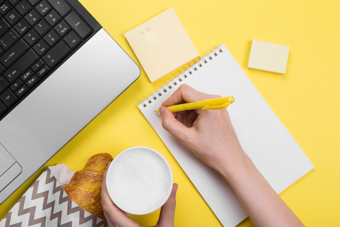 Working morning. Girl works for laptop. Breakfast at work - cappuccino, croissant, notebook on yellow background. Top view